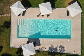 Luchtbeeld van een rechthoekig zwembad met helderblauw water, omringd door een betegeld zonnedek en vier witte parasols.