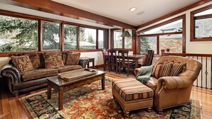 A spacious living room with a brown leather sofa, a wooden coffee table, and a striped ottoman. There is a dining table with chairs and a chandelier. Large windows provide a view of trees and a stone wall.