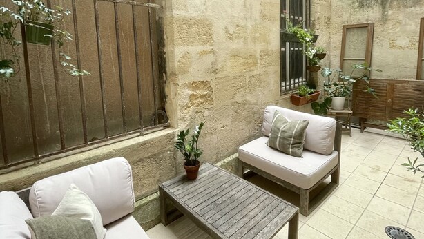 A cozy courtyard with a stone wall, a wooden bench, a cushioned sofa, and potted plants. - Charming Artistic and Historic Chartrons Flat with Private Patio
 (Bordeaux)