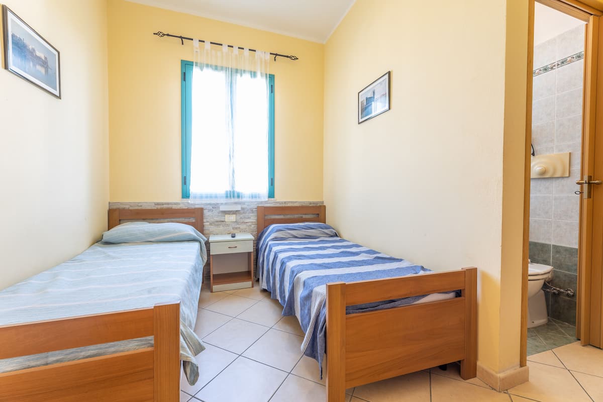 Una habitación pequeña y luminosa con dos camas de madera, una ventana con cortinas, una mesita de noche y una puerta que da al baño.