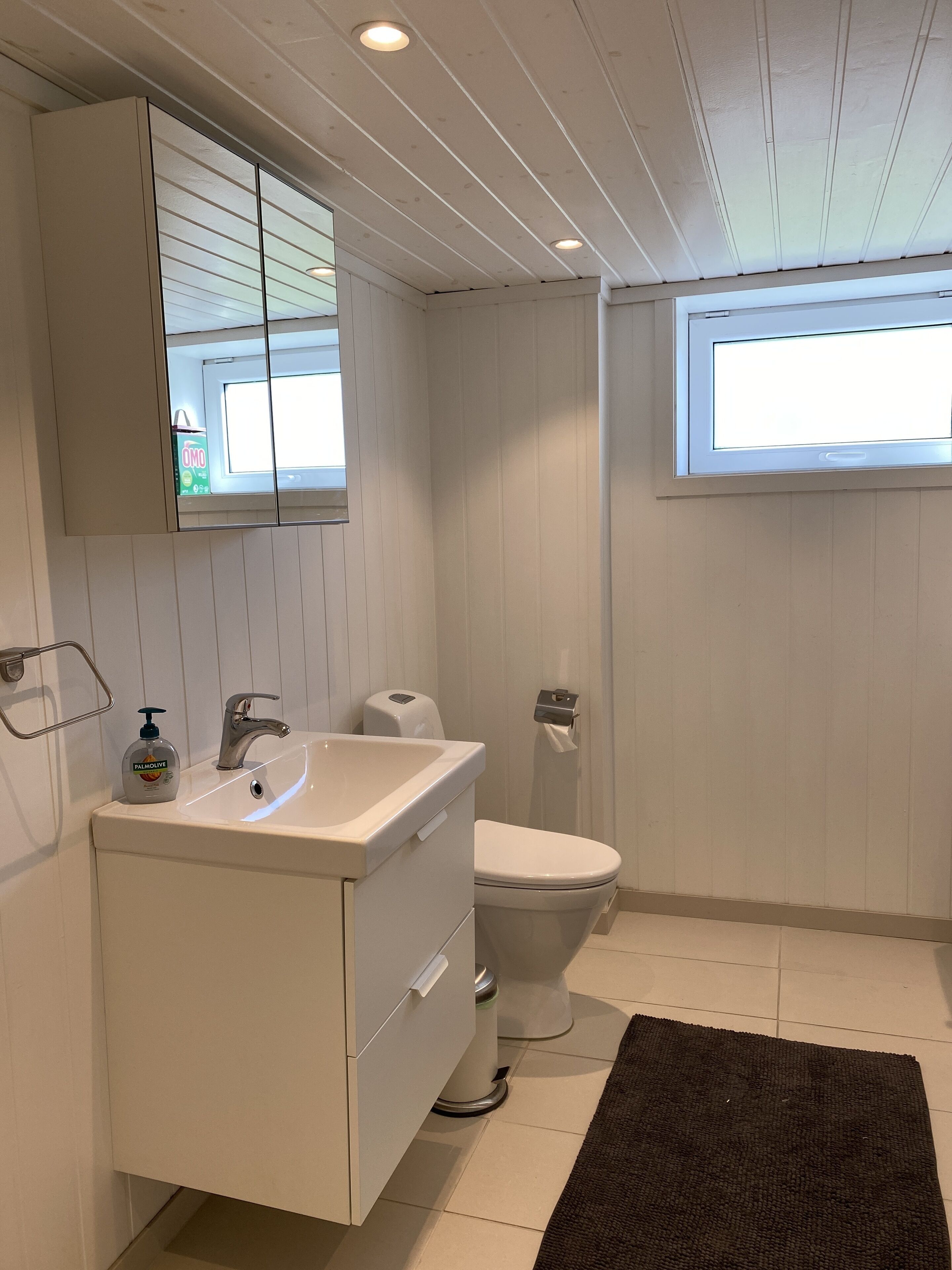 Ein Badezimmer mit Waschbecken, Spiegel und Toilette. Über der Toilette befindet sich ein Fenster und auf dem Boden ein dunkler Teppich.