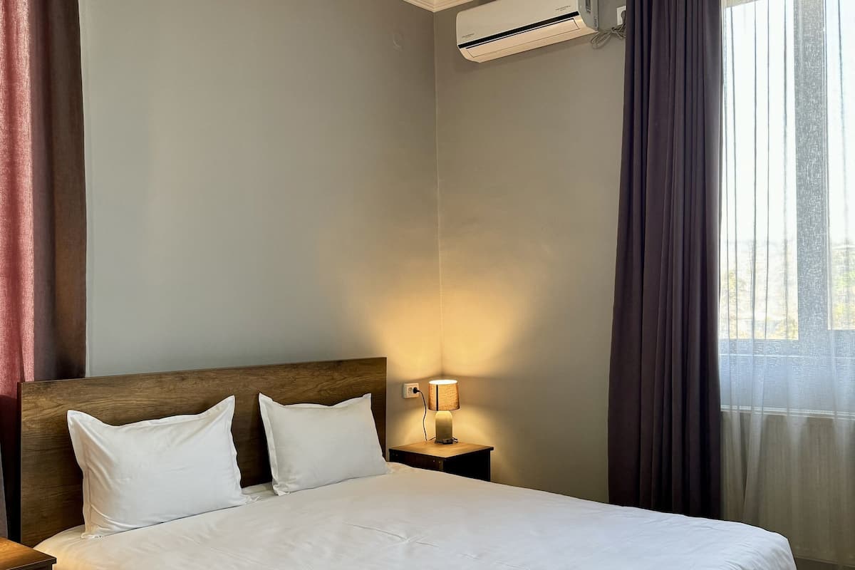 Une chambre d’hôtel avec un grand lit, une table de chevet avec une lampe, un appareil de climatisation et une fenêtre avec des rideaux.