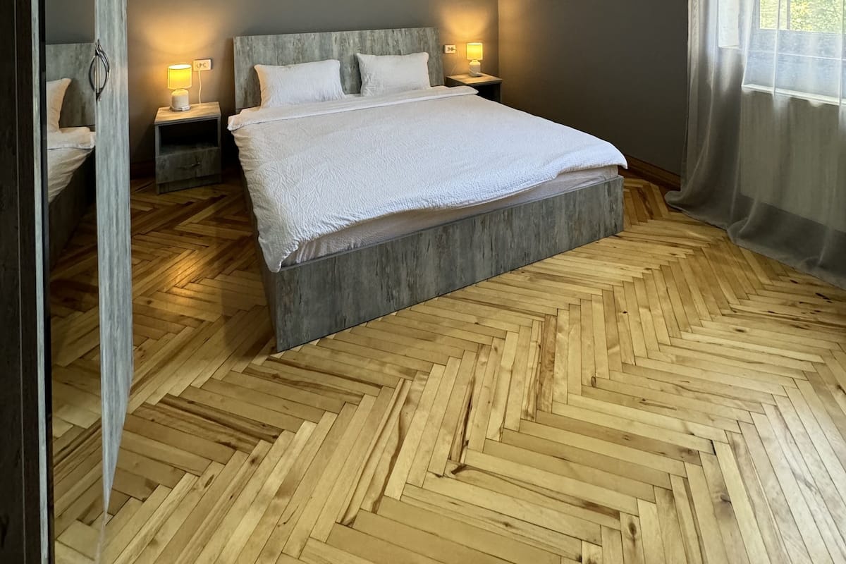 Une chambre moderne avec un grand lit, un parquet en bois disposé en point de Hongrie et une vue sur la verdure par la fenêtre.