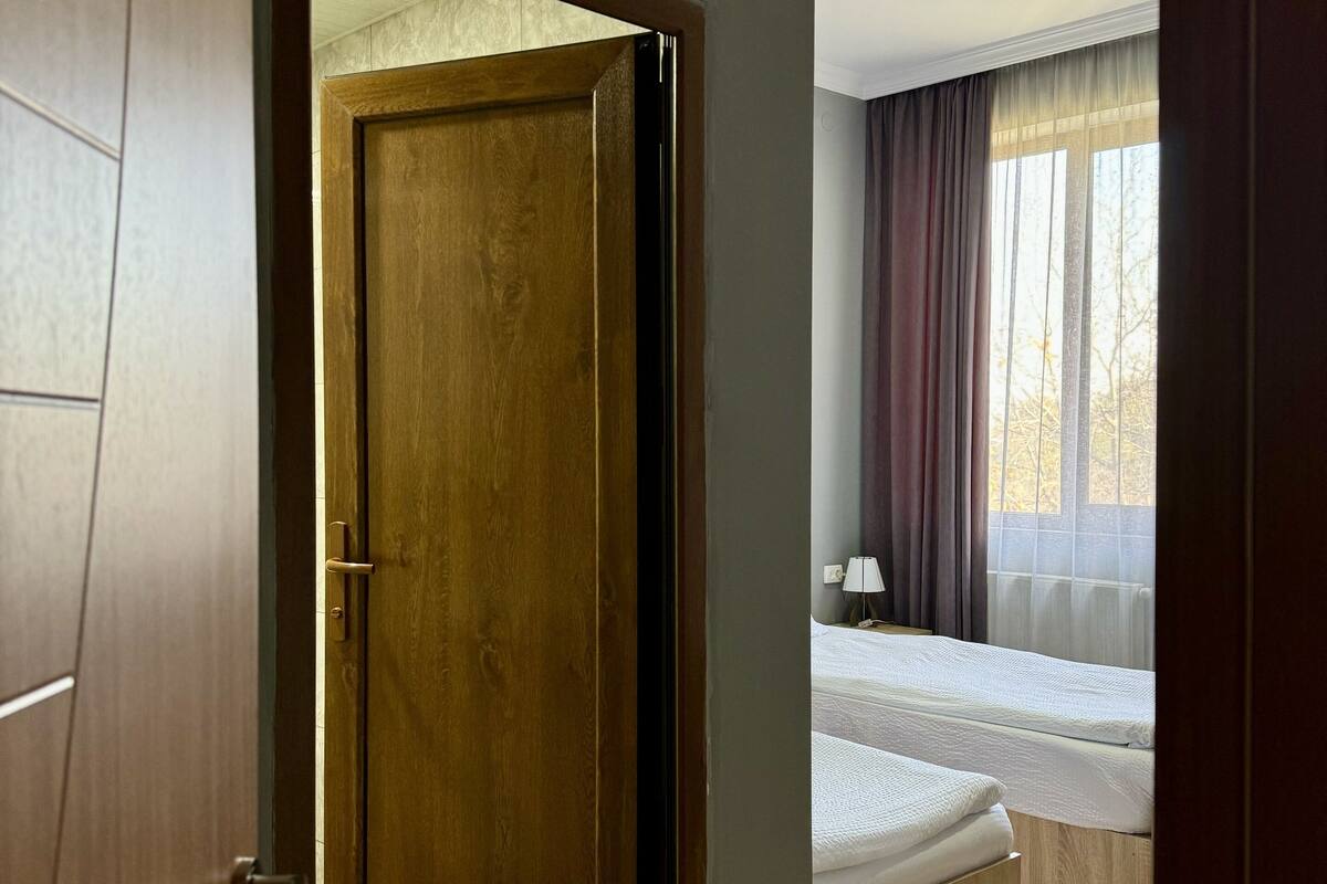 Une chambre d’hôtel avec une porte en bois, un lit avec du linge de lit blanc, une table de chevet avec une lampe et une fenêtre avec des rideaux.