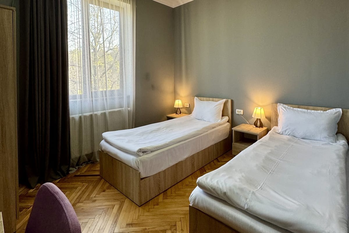 Une chambre d’hôtel avec deux lits simples, un plancher en bois, une fenêtre avec des rideaux et un bureau avec une chaise.