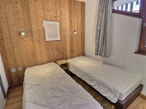 Quarto com duas camas de solteiro, paredes de painel de madeira, um quadro emoldurado, uma mesinha e uma janela com cortinas.