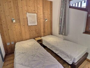 Quarto com duas camas de solteiro, paredes de painel de madeira, um quadro emoldurado, uma mesinha e uma janela com cortinas.