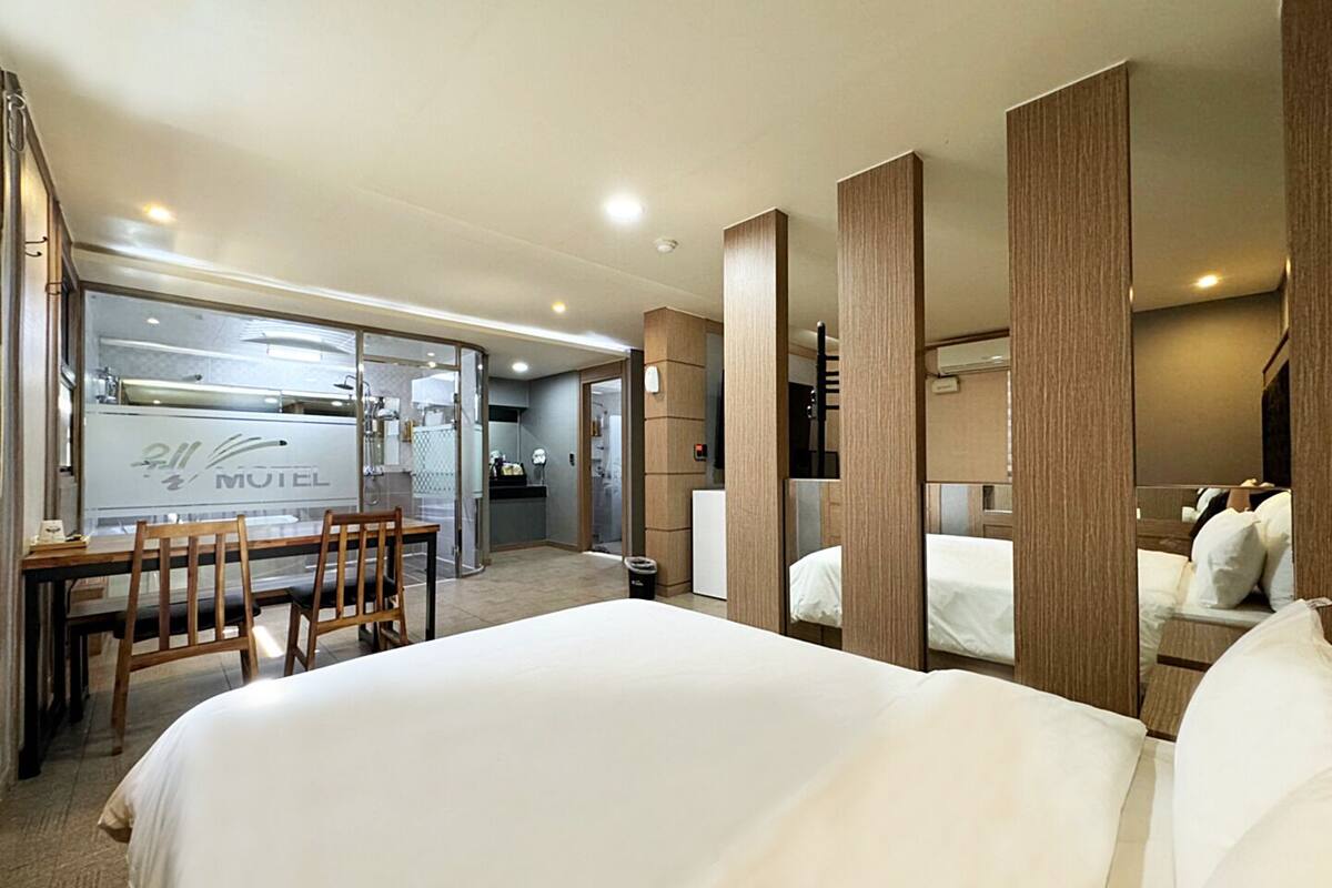 A hotel room with a bed, a desk, and a chair. There is a kitchenette in the background.