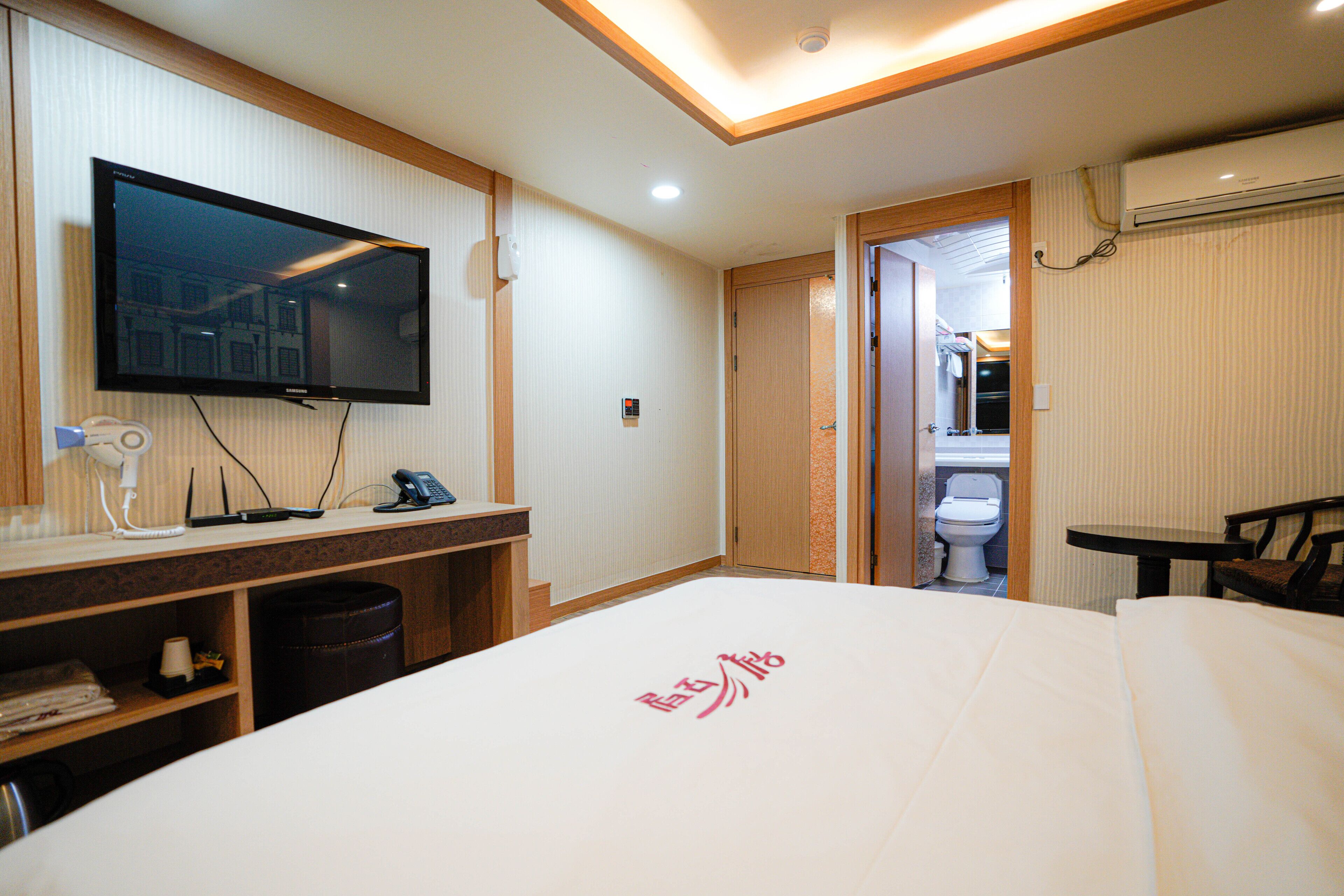 A hotel room with a bed, a TV, a desk with a phone, a trash bin, and a bathroom visible through an open door.