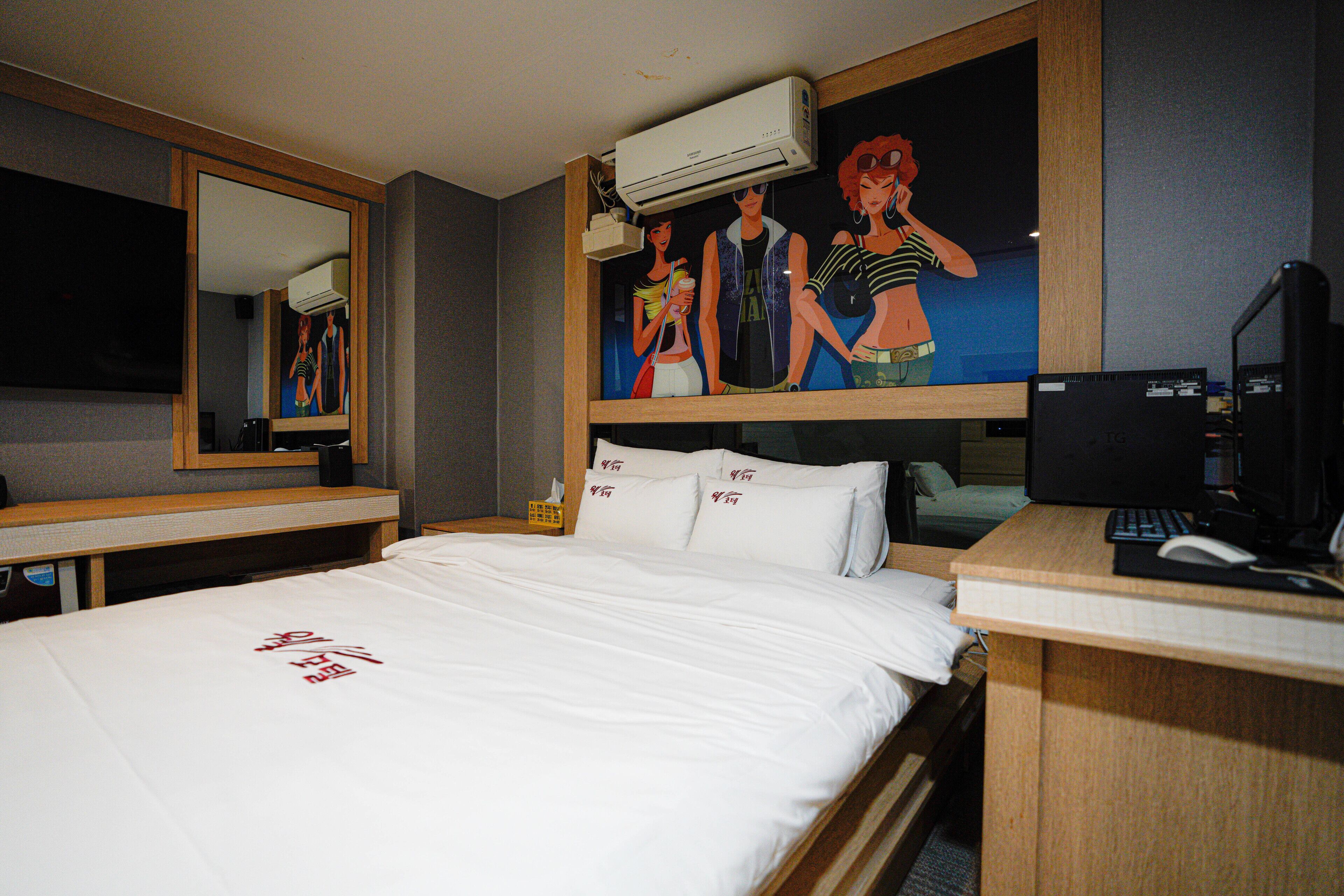 Habitación de hotel con cama, escritorio con computadora, espejo y un cuadro en la pared.