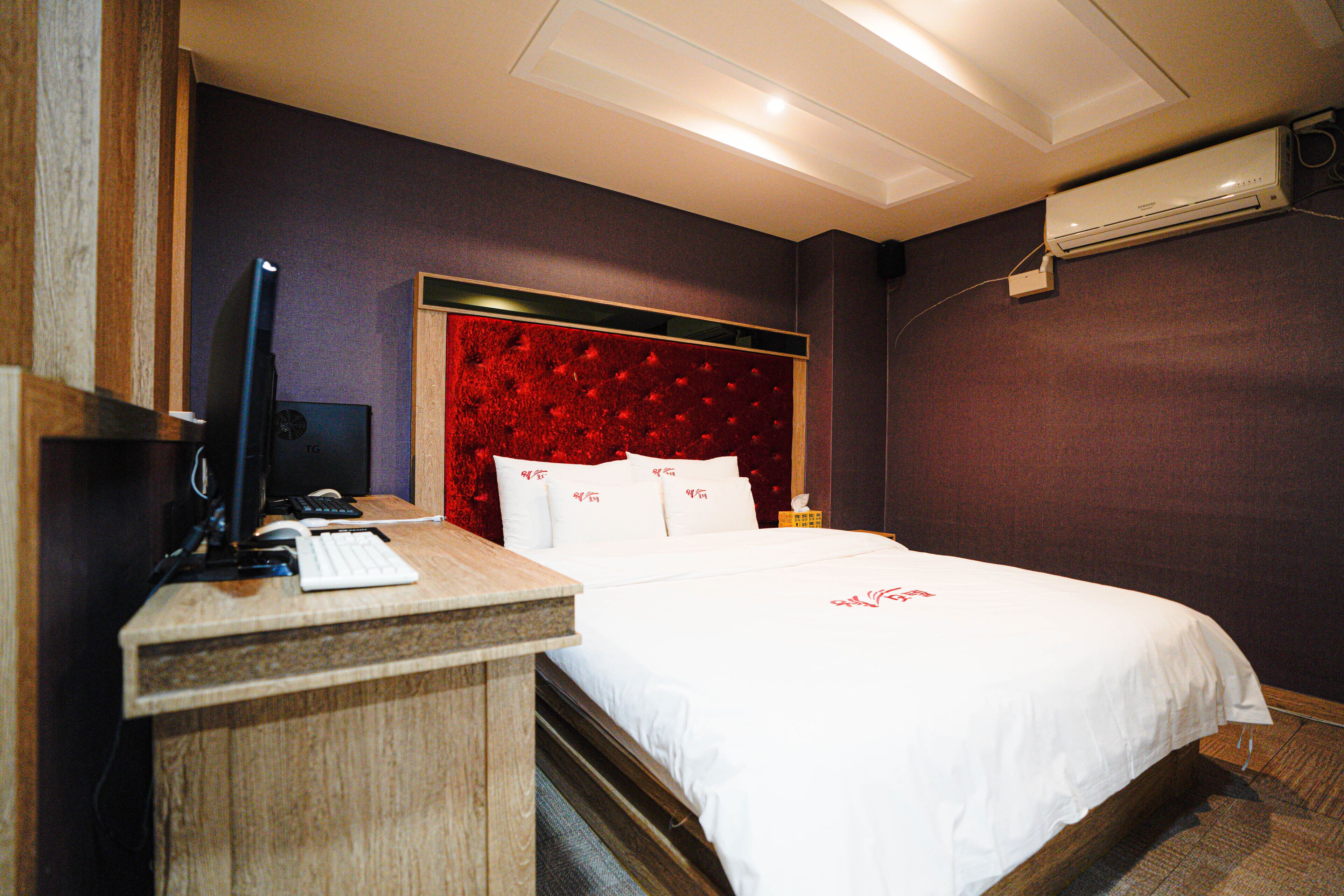 Habitación de hotel con un escritorio de madera, una cama con ropa blanca, un cabecero tapizado en rojo y un equipo de aire acondicionado en la pared.