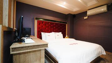 Habitación de hotel con un escritorio de madera, una cama con ropa blanca, un cabecero tapizado en rojo y un equipo de aire acondicionado en la pared.