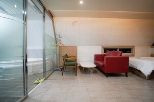 Una habitación de hotel moderna con cabina de ducha de vidrio, un sofá rojo, una mesita redonda pequeña y una cama con cabecera blanca.