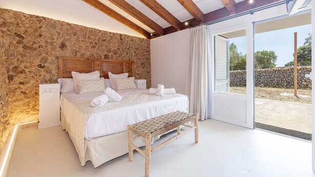 Ein Schlafzimmer mit Steinwand, Holzbalkendecke, einem Bett mit weißer Bettwäsche, einer Bank und Blick ins Freie.