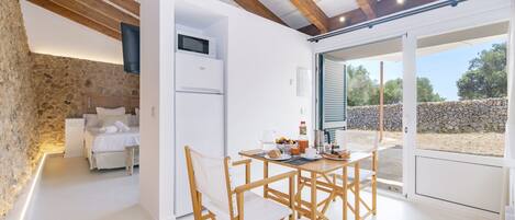 A modern room with a stone wall, a bed, a white refrigerator, a dining table with chairs, and a large sliding glass door leading to an outdoor area.