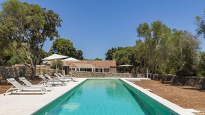 Ein rechteckiger Swimmingpool mit einer Holzterrasse, umgeben von Liegestühlen und Sonnenschirmen, mit einer Steinmauer und Bäumen im Hintergrund.