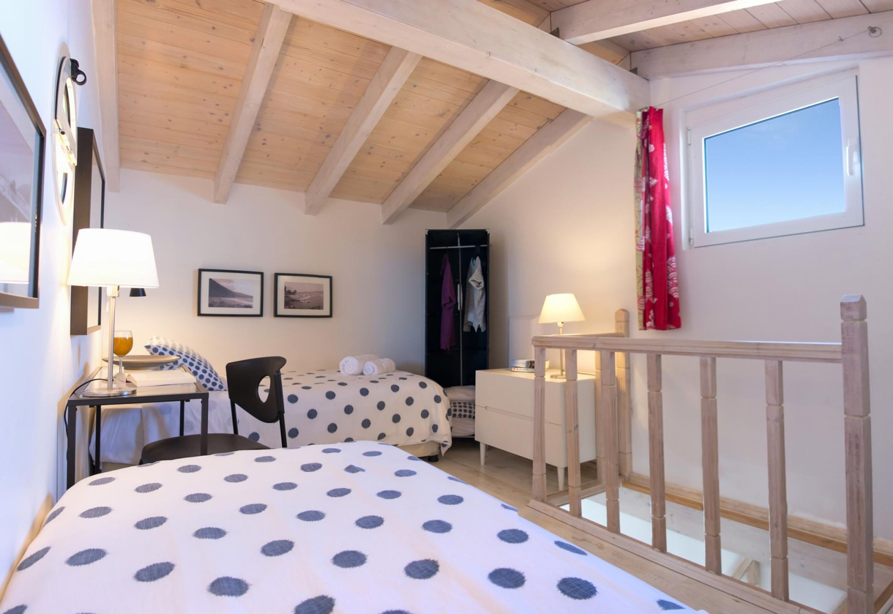 A bedroom with a wooden ceiling, two beds with polka dot bedding, a desk with a lamp, a chair, a wardrobe, and a window with a red curtain.