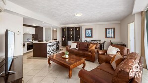 Ein modernes Wohnzimmer mit einer braunen Ledersofa, einem Couchtisch aus Holz und einem Fernseher auf einem Standfuß. Im Hintergrund ist eine Küche mit weißen Schränken und ein Essbereich mit Tisch und Stühlen zu sehen.