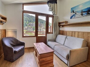 A room with a sofa, a chair, a wooden table, a window with a view of buildings and trees, and a painting on the wall.