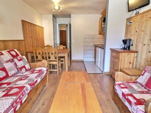 A cozy living room with a sofa, a dining table, and chairs, a kitchen with a microwave and a coffee machine, and wooden walls.