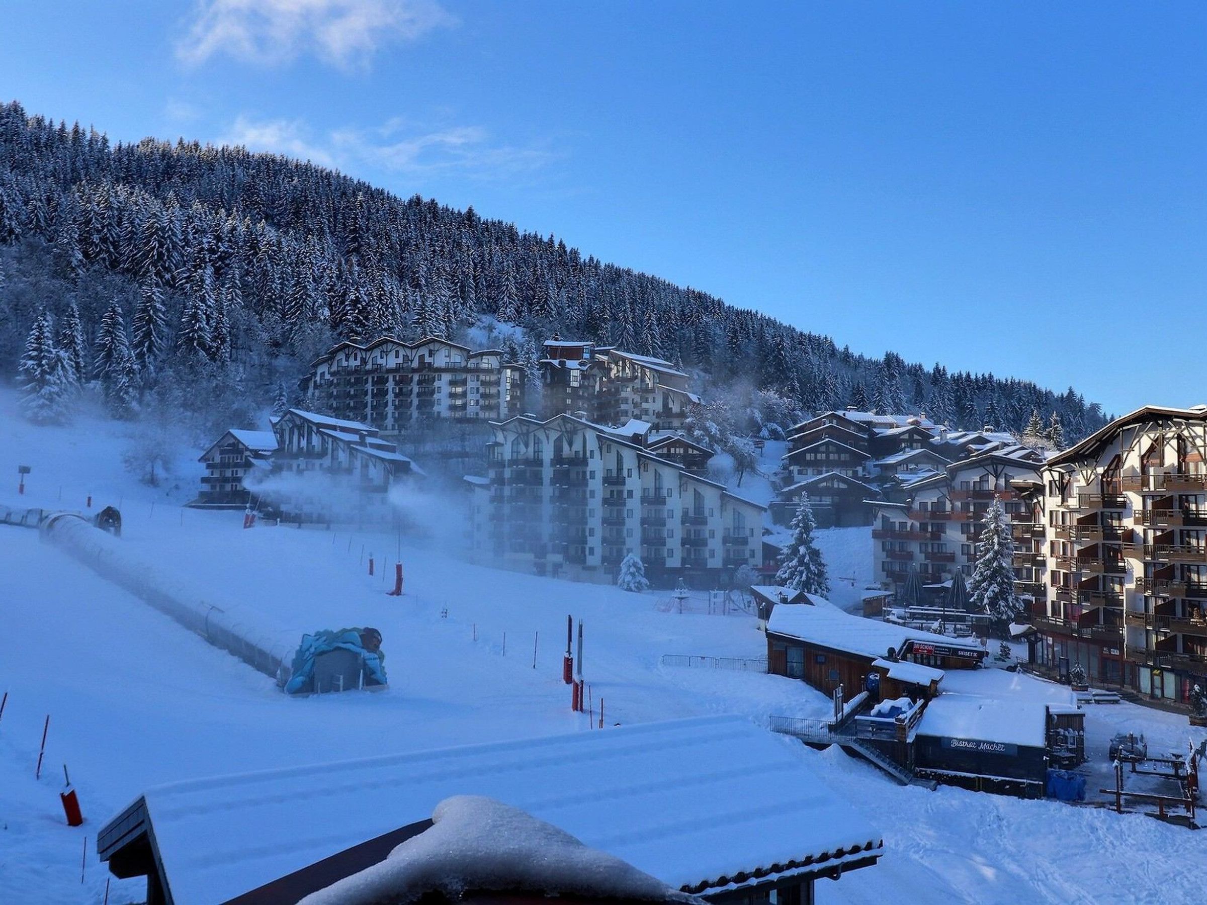 A snow-covered ski resort with multiple buildings, including a hotel with balconies and a restaurant.