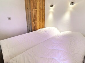 A bedroom with a wooden door, a bed with white bedding, and two wall-mounted light fixtures. - Cozy 2-Room, Ski-in/Ski-out, Near Shops (La Tania)