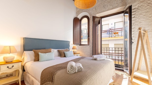 A bedroom with a large bed, two bedside tables with lamps, a wooden floor, a balcony with a wrought iron railing, and a stone wall. - Triana Acacias by Valcambre  (Seville)