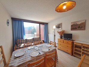 Ein Essbereich mit einem Tisch für vier Personen, einem Fernseher auf einem Holzschrank und einem Fenster mit Vorhängen.