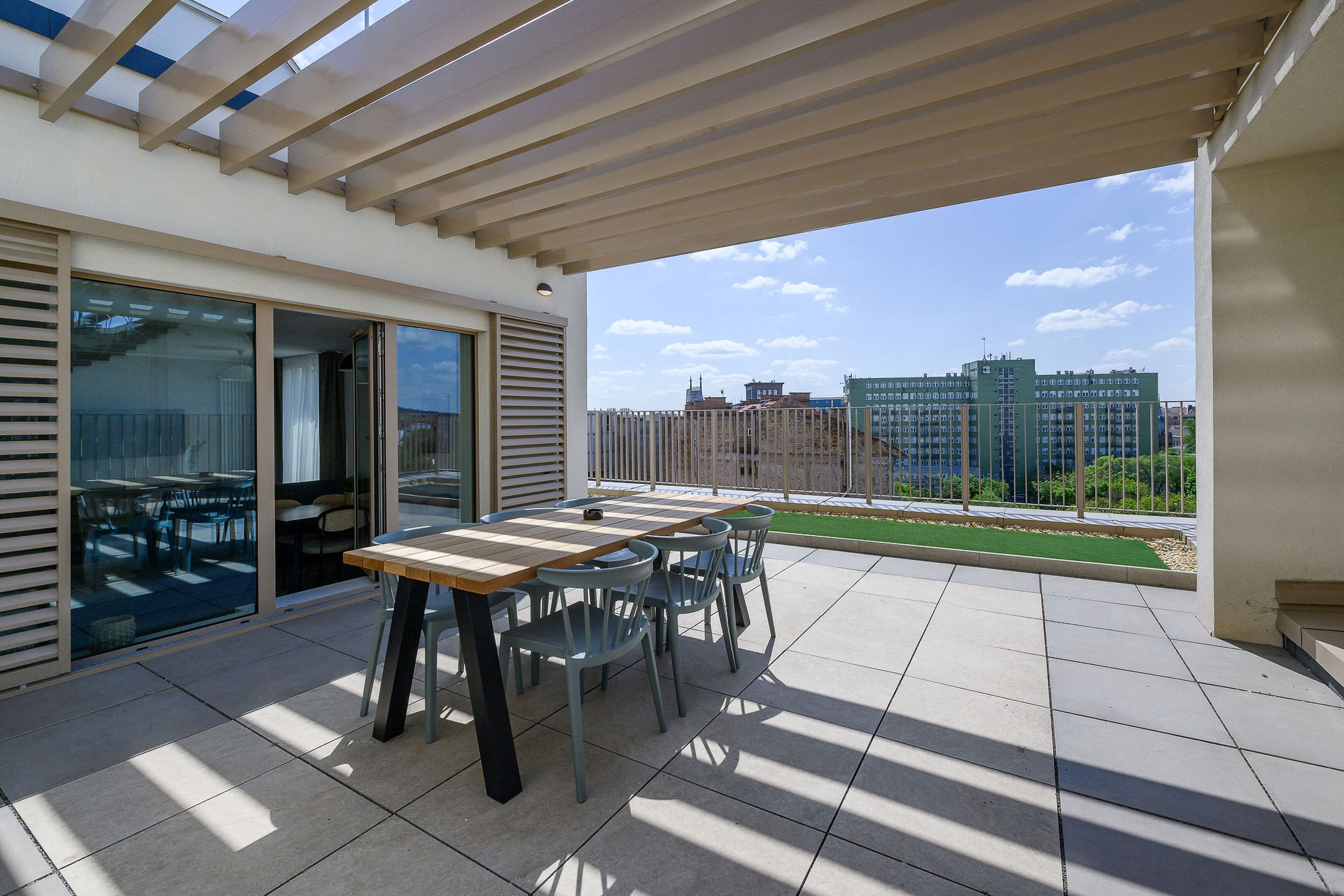 A covered patio with a table and chairs, large sliding glass doors, and a view of buildings in the background.