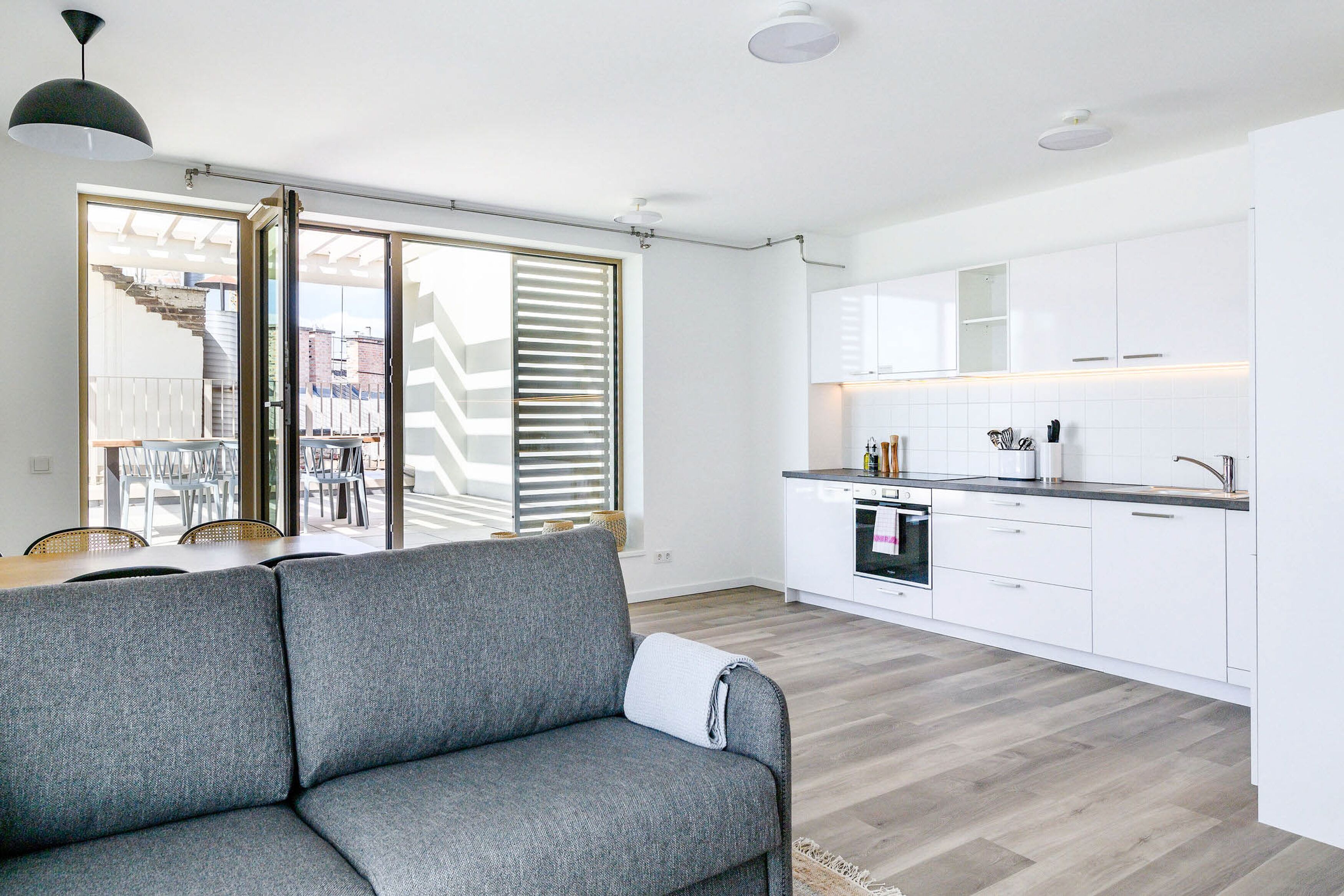 A modern apartment with a grey sofa, a dining area with a table and chairs, a kitchen with white cabinets, and a large sliding glass door leading to a balcony.