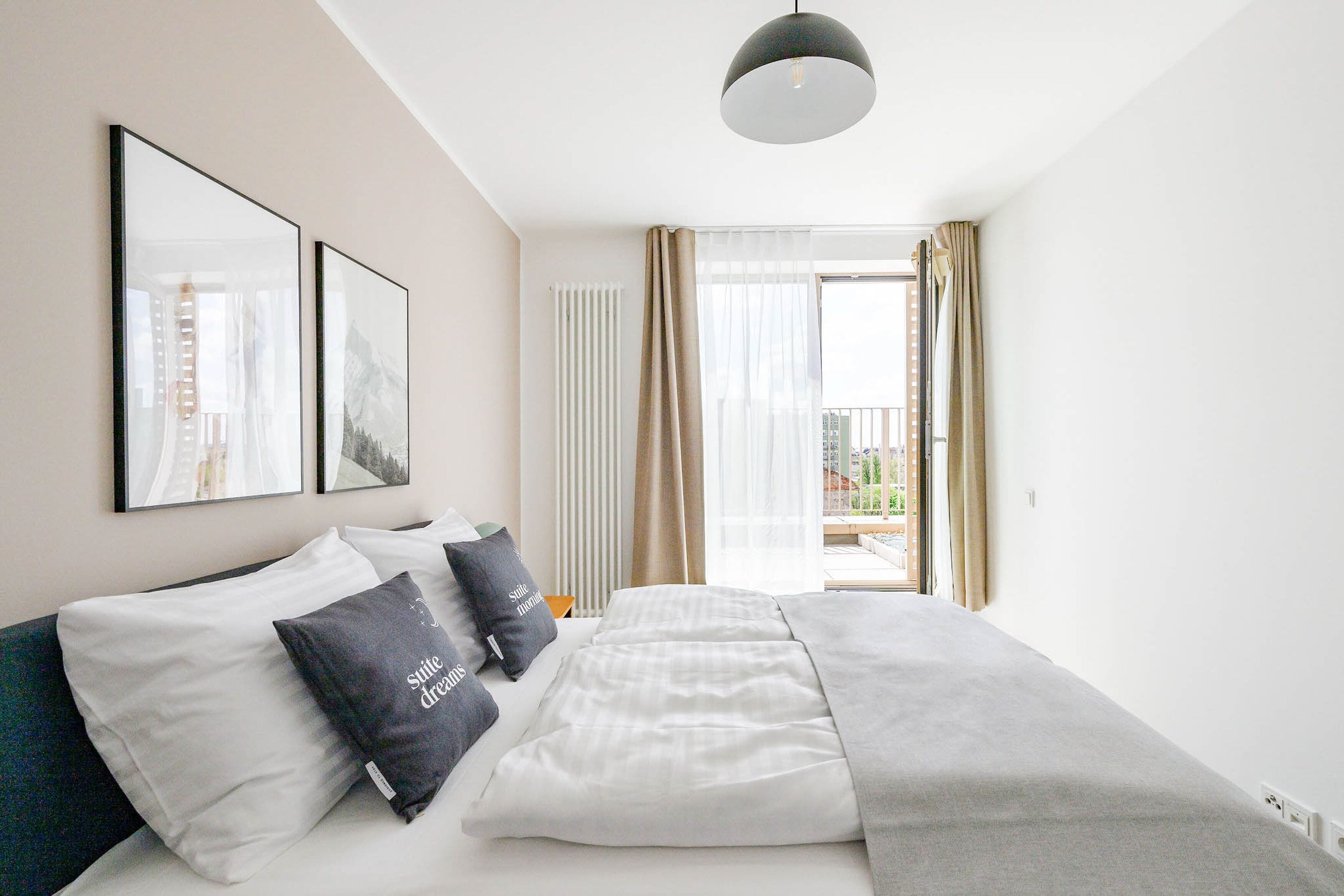 A modern hotel room with a large bed, two pillows, a bedside table, a hanging light fixture, and a view of the city through a large window.
