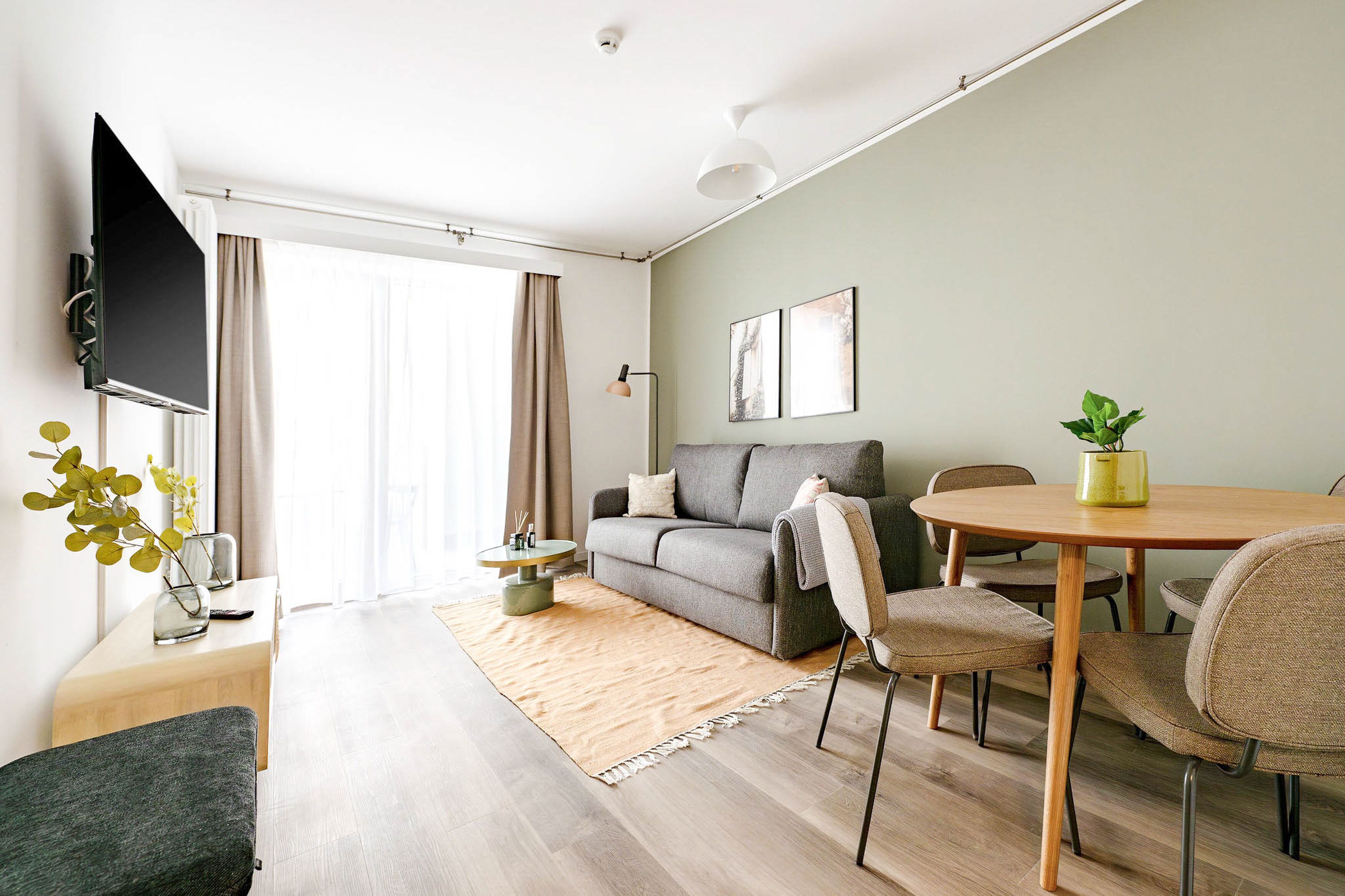 A modern living room with a sofa, a dining table, and chairs. There is a TV mounted on the wall, a plant on the table, and a window with curtains.