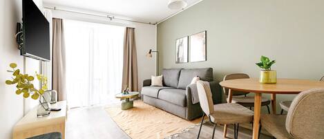 A modern living room with a sofa, a dining table, and chairs. There is a TV mounted on the wall, a plant on the table, and a window with curtains.