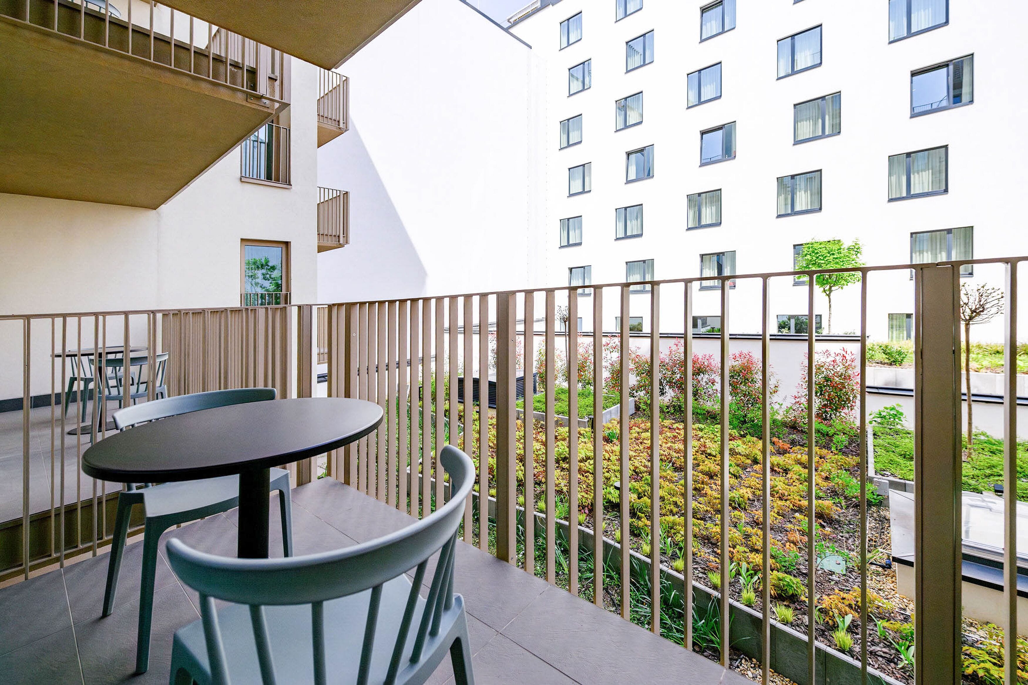 Uma varanda com mesa e cadeiras, vista de um edifício com várias janelas e um jardim com plantas e uma árvore.