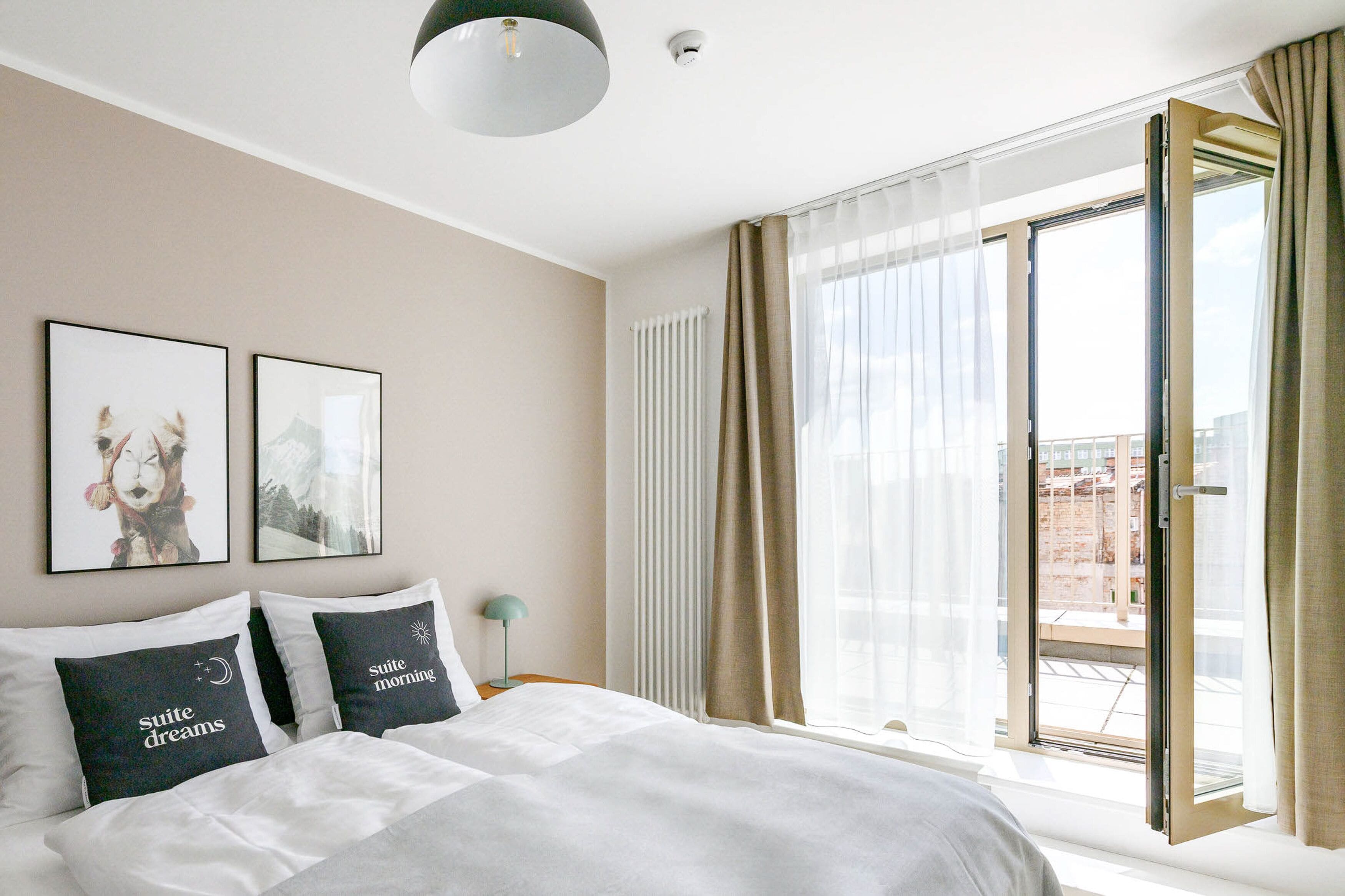 Et hotelværelse med en seng, to puder med tekst, et natbord med en lampe, et vindue med gardiner og udsigt til bygninger udenfor.