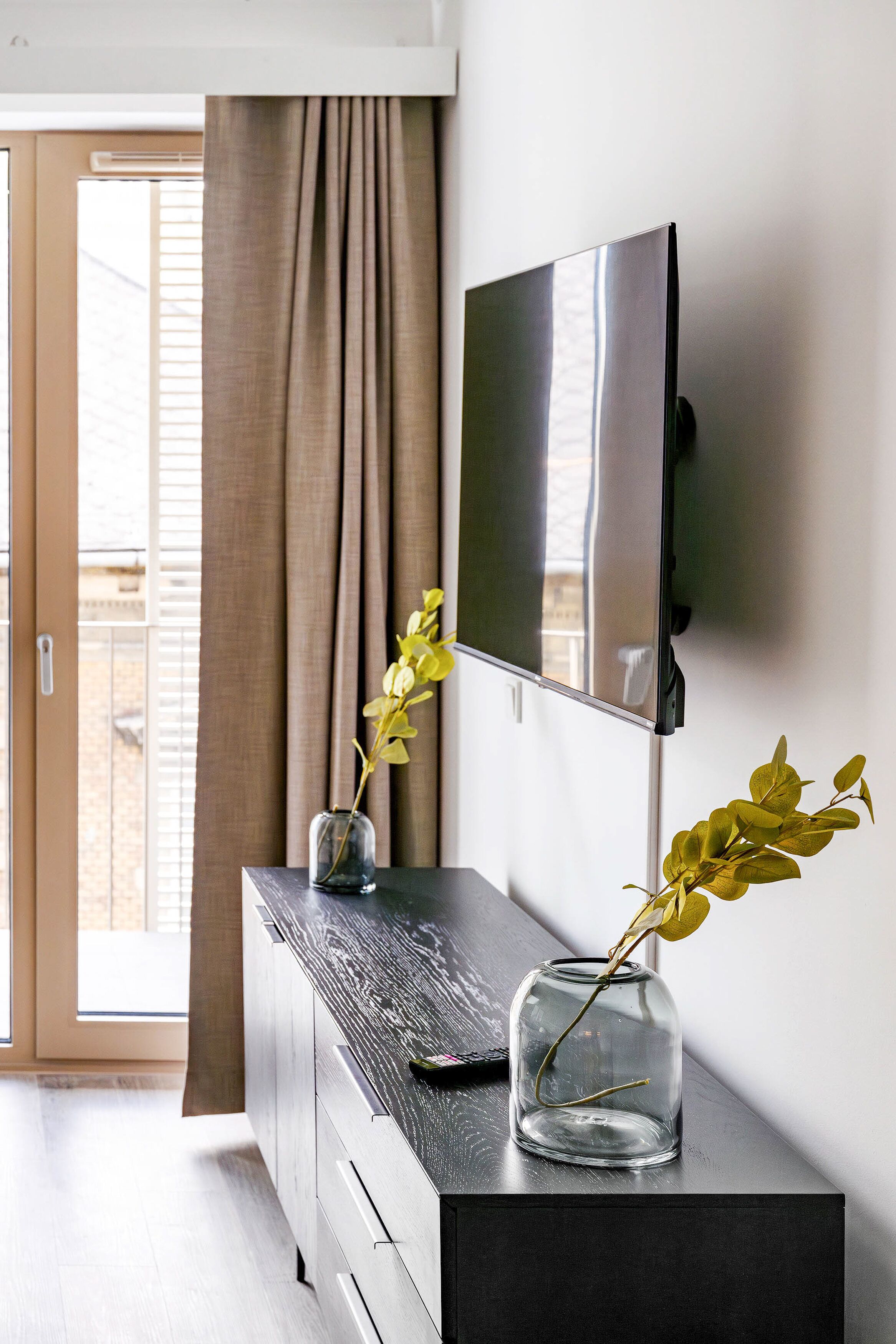 A modern living room with a flat-screen TV mounted on the wall, a dark wooden console table, and a glass vase with a plant.