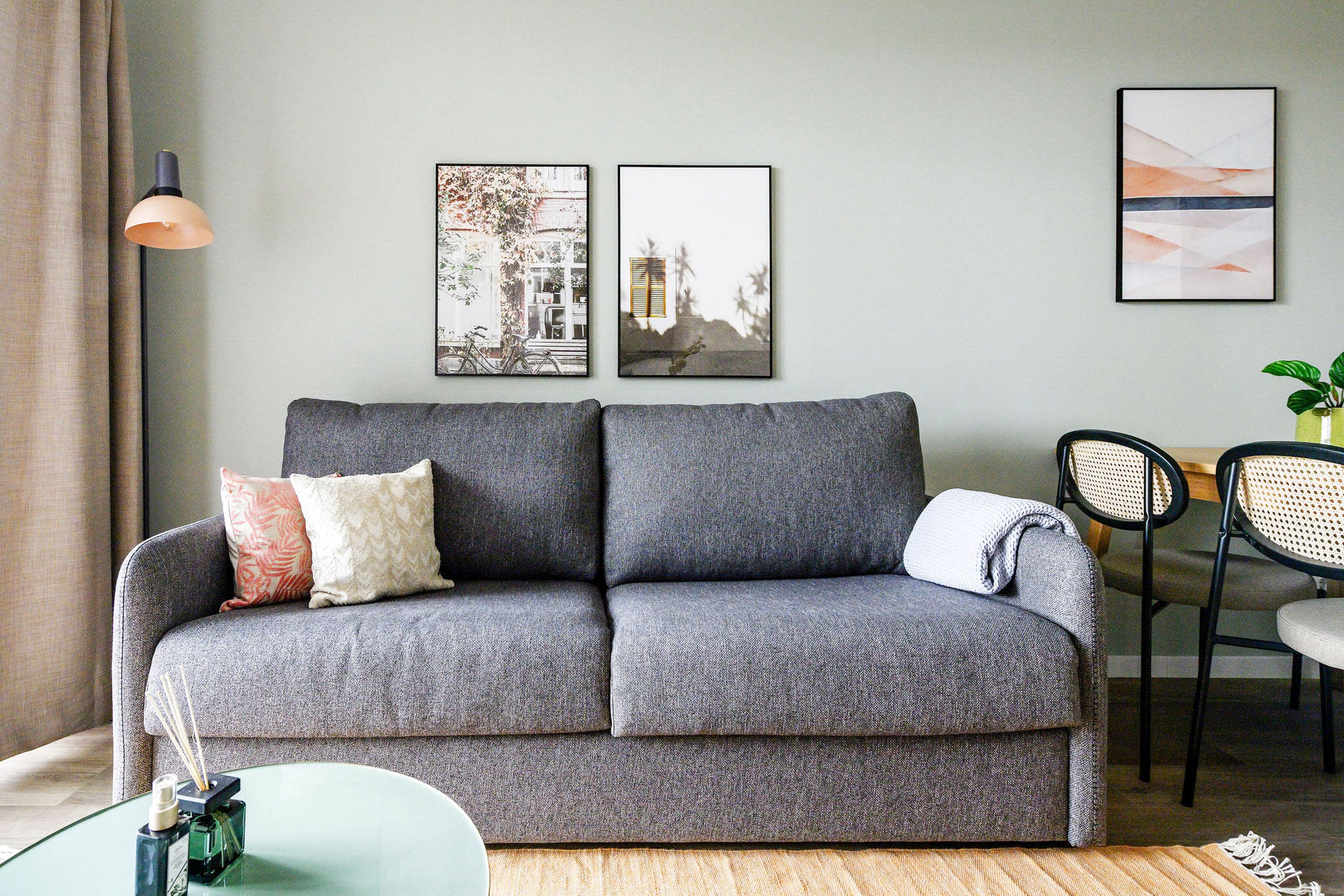 Ein graues Sofa mit Kissen, ein Glastisch mit einer Flasche und einer Kerze, sowie zwei gerahmte Bilder an der Wand.