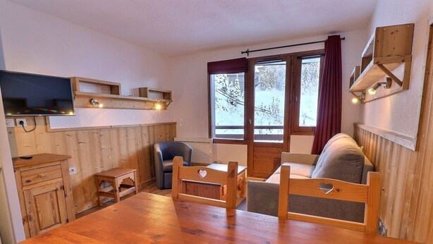 Une pièce chaleureuse avec une table à manger en bois, un canapé, un téléviseur fixé au mur et une fenêtre donnant sur des arbres couverts de neige.