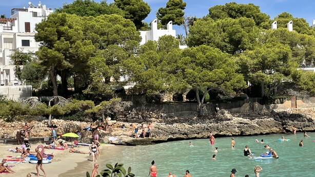 Ein Strand mit badenden und sonnenbadenden Menschen, umgeben von Bäumen und Gebäuden.