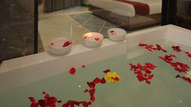 A modern hotel room with a large bed, a glass-enclosed bathtub with rose petals, and a view of the room's interior through a mirror. - Hotel Taj Crown Plaza (Agra)