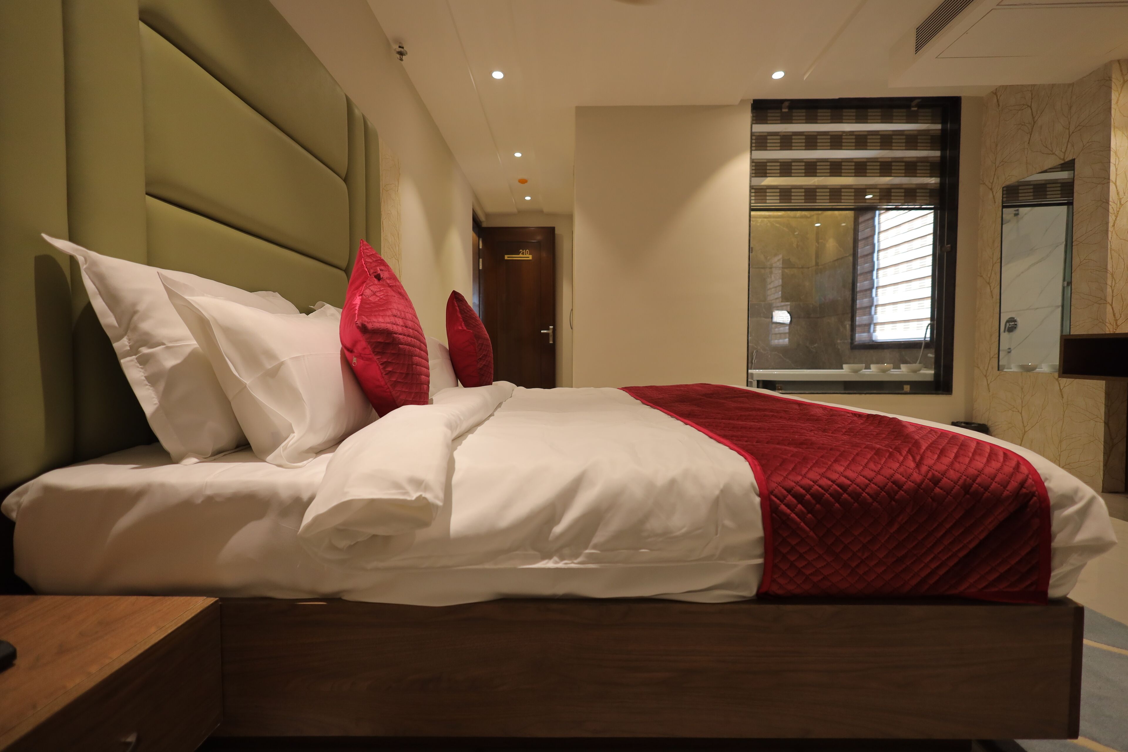 A modern hotel room with a large bed, a wooden nightstand, a red and white bedspread, and a large window with blinds.