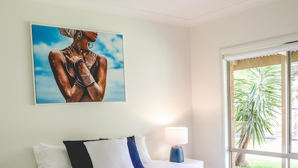 A bedroom with a bed, a ceiling fan, a window with a view of greenery, and a painting on the wall.