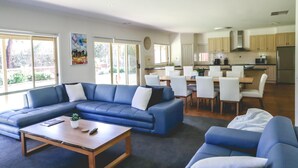 A modern living room with a blue sectional sofa, a wooden coffee table, and a dining area with white chairs and a kitchen in the background.