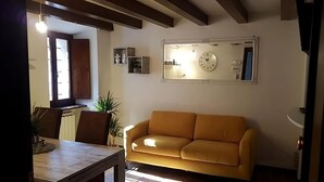 Ein Wohnzimmer mit einem gelben Sofa, einem Esstisch mit Stühlen, einer Topfpflanze und einer Wanduhr.