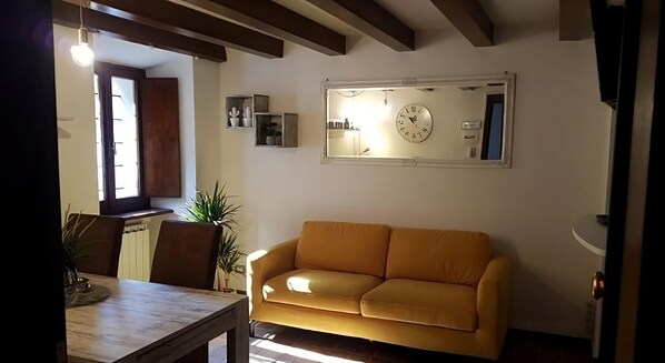 A living room with a yellow sofa, a dining table with chairs, a potted plant, and a wall clock.