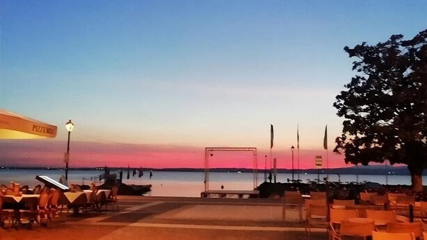 Ein Sitzbereich im Freien mit Tischen und Stühlen, der auf ein Gewässer im Sonnenuntergang blickt.