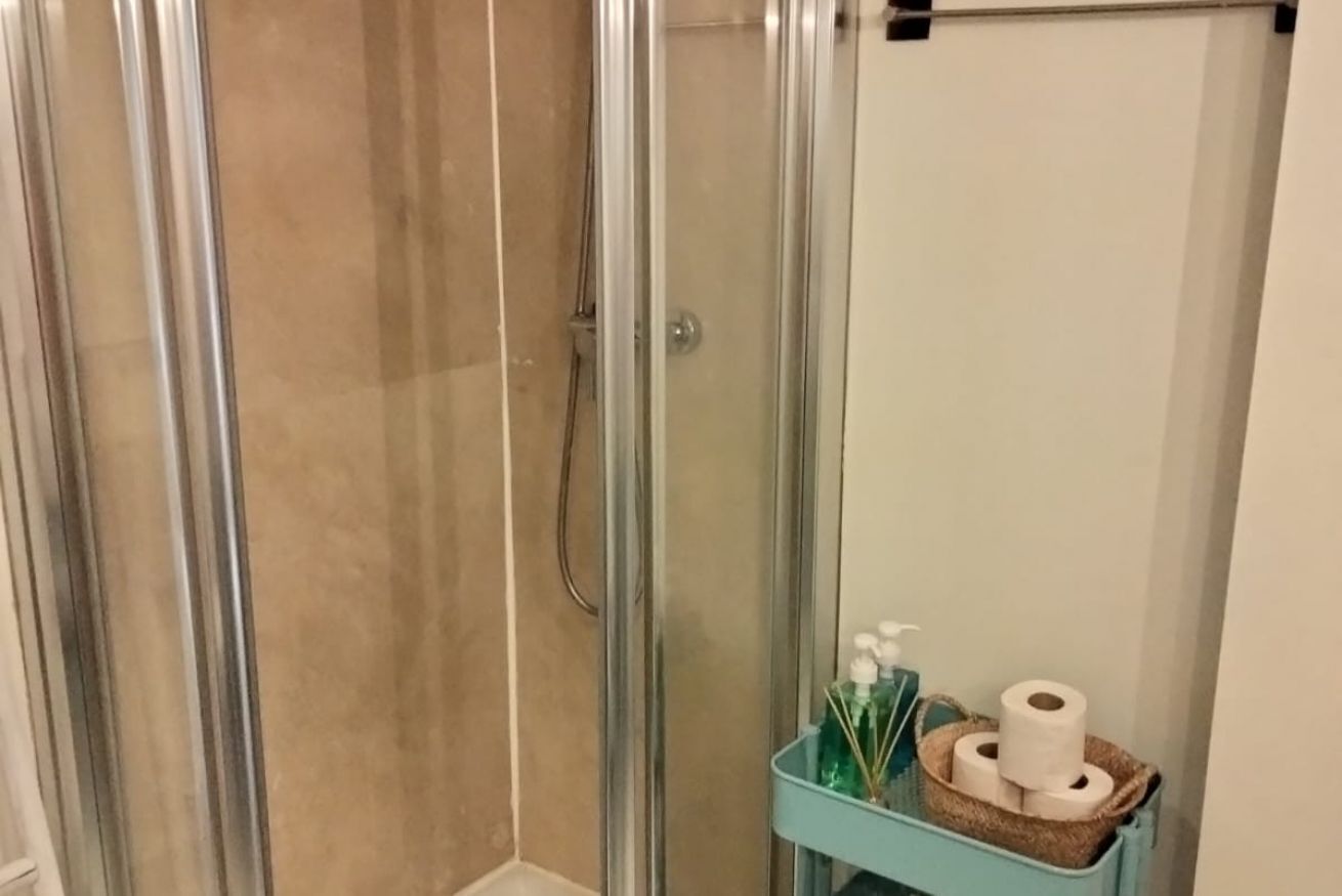 Ein durch eine Glaswand abgetrennter Duschbereich mit einer beige gefliesten Wand, einem Handtuchhalter und einer kleinen Ablage für Toilettenartikel.