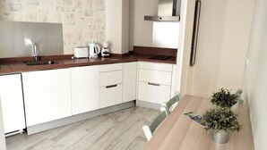 Une cuisine moderne avec des meubles blancs, un évier en acier inoxydable, un grille-pain et une machine à café. On y trouve une table à manger en bois, des chaises et, à droite, un pot de fleurs.