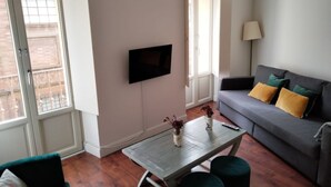 Un salon avec un canapé gris, un téléviseur à écran plat fixé au mur, une petite table sur laquelle sont posés deux vases et une lampe.