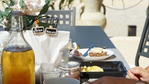 Una mesa dispuesta con alimentos para el desayuno, como cruasanes, una botella de jugo de naranja y un vaso de agua, con vistas a palmeras y un edificio al fondo.